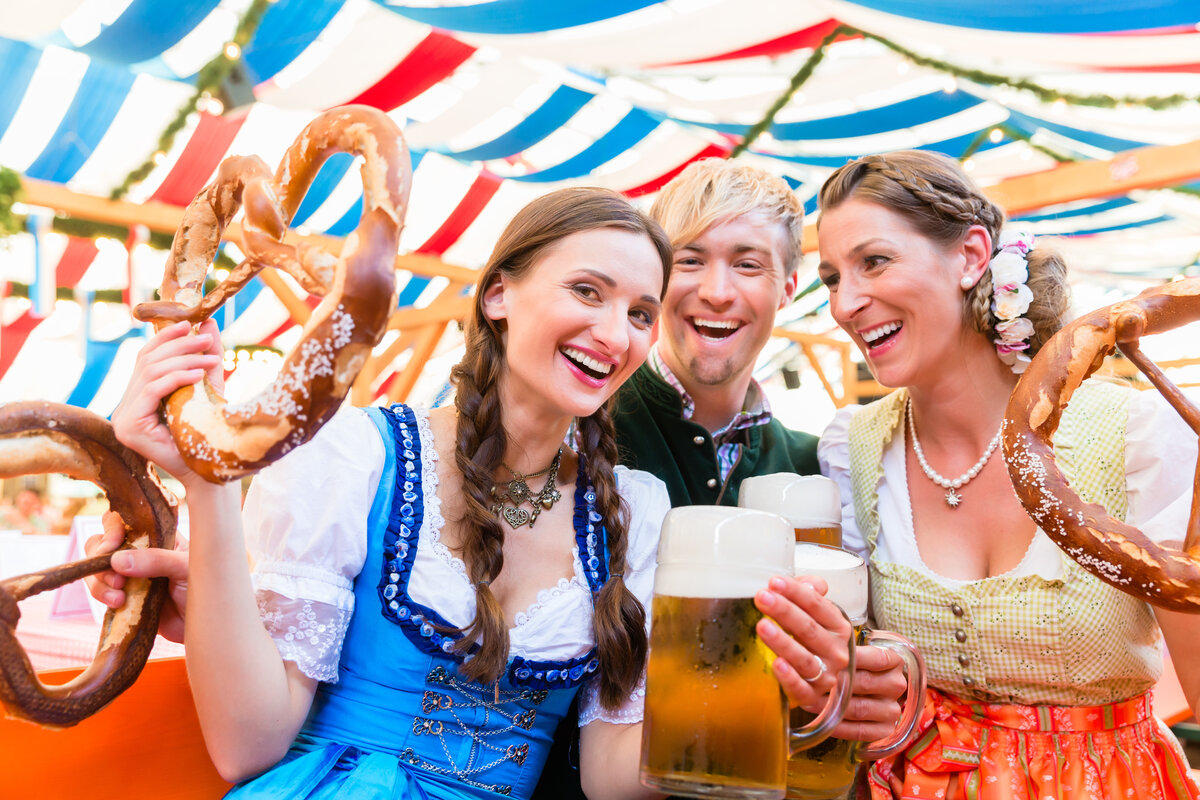 Two women and a man in traditional German clothes at Oktoberfest enjoying large steins of beer and huge pretzels.